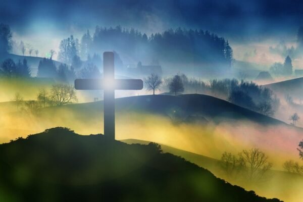 Silhouette of a cross on a hill with a misty, forested landscape in the background and a bright light shining through the cross at sunrise or sunset.