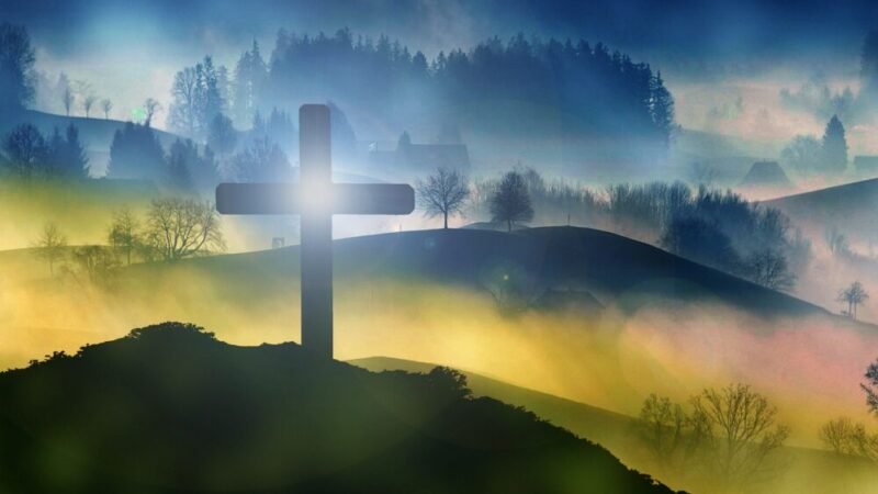Silhouette of a cross on a hill with a misty, forested landscape in the background and a bright light shining through the cross at sunrise or sunset.
