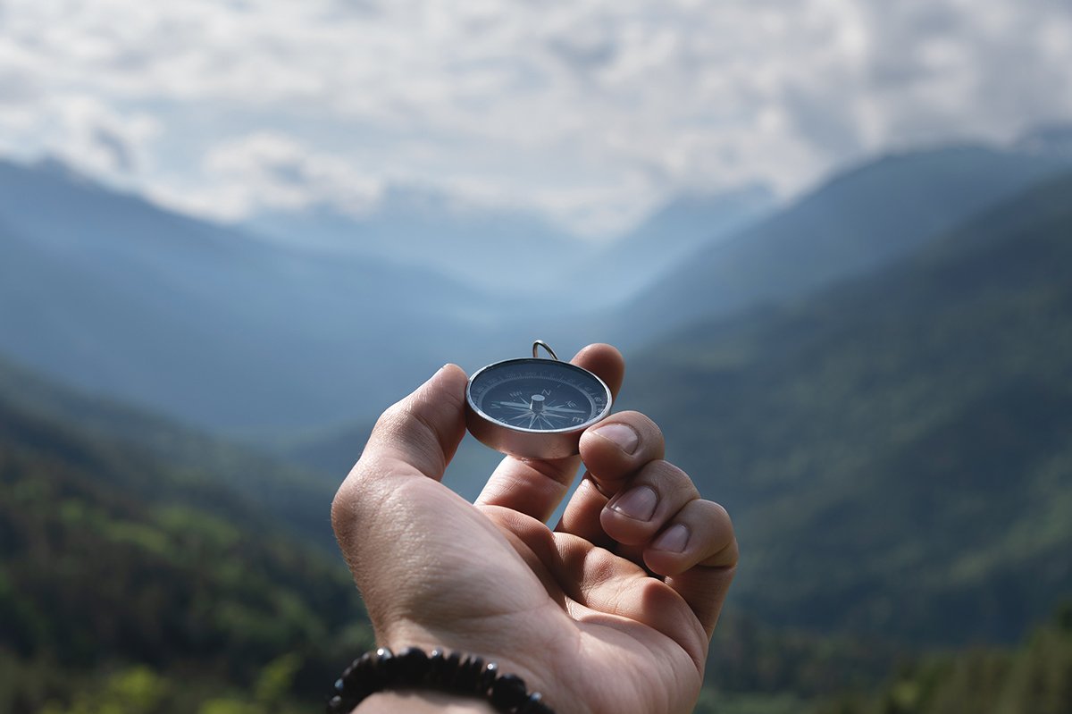 Magnetic compass in the palm of a male hand against the backdrop of a mountain range in the clouds in the summer outdoors, travel, first person view