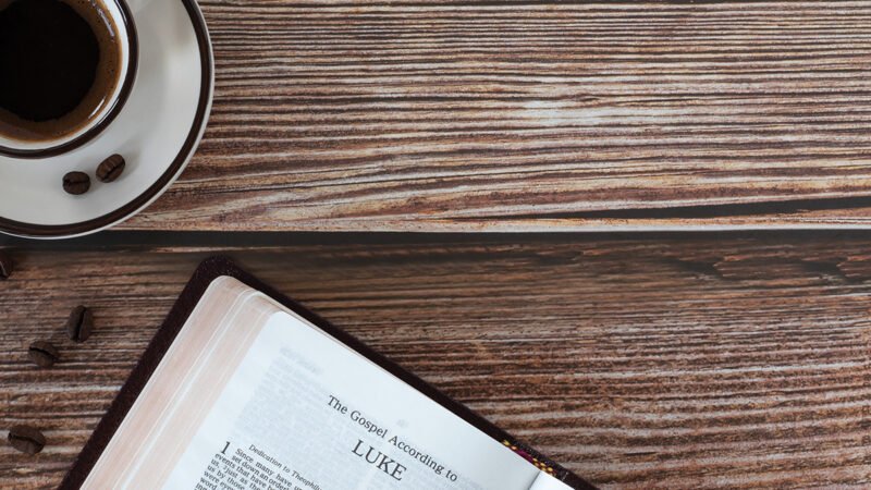 Open holy bible book Luke gospel with coffee cup on wooden background, copy space, top view